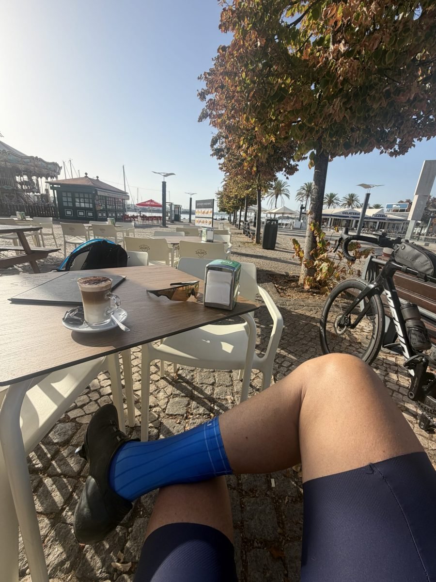 Coffee on a riverside café table in Portimão, legs stretched out in blue socks, road bike leaning against a tree with autumn leaves