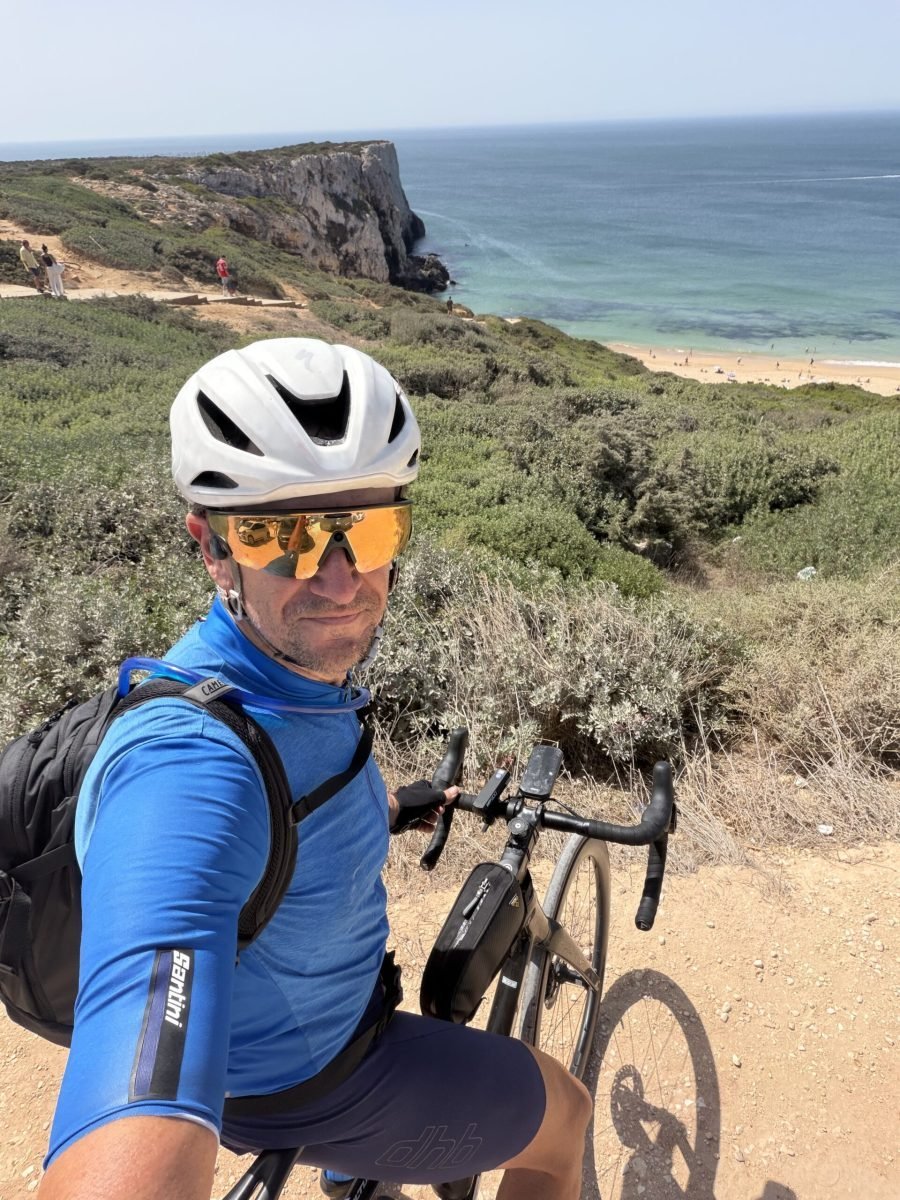 Selfie of Richard in a blue jersey, white helmet and orange-lensed glasses, with his bike, scrubby clifftop and the Atlantic coast behind