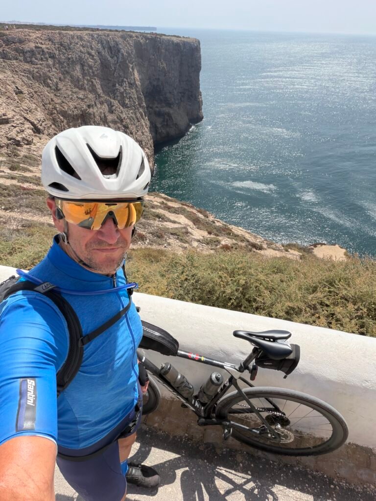 Selfie of Richard at Cabo de São Vicente in a blue jersey and white helmet, sheer cliffs and the Atlantic to his left, his bike leaning on the wall to his right