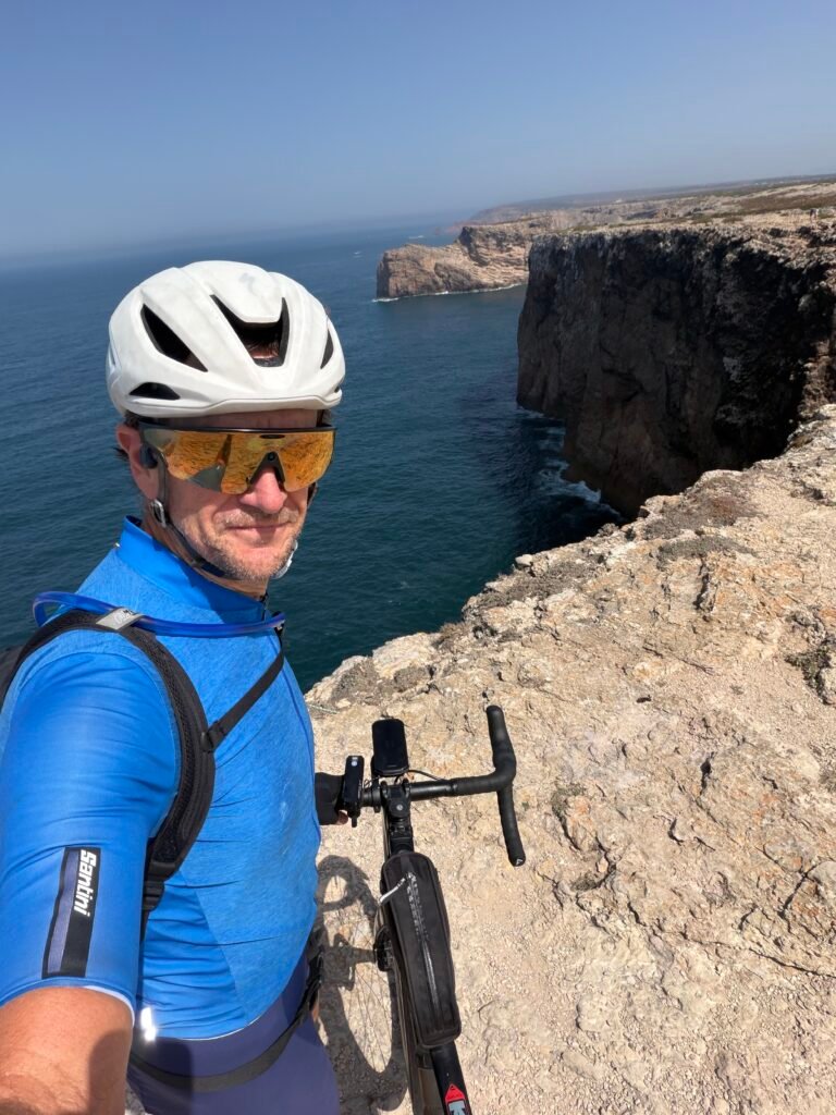 Selfie of Richard at Cabo de São Vicente in a blue jersey and white helmet, with his bike beside him and sheer cliffs above the Atlantic to his right
