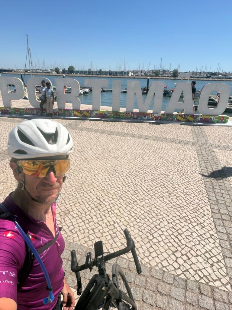 Richard in a purple Maglia Ciclamino jersey and white helmet taking a selfie in front of the big white PORTIMÃO letters on the waterfront, with the marina and boats behind