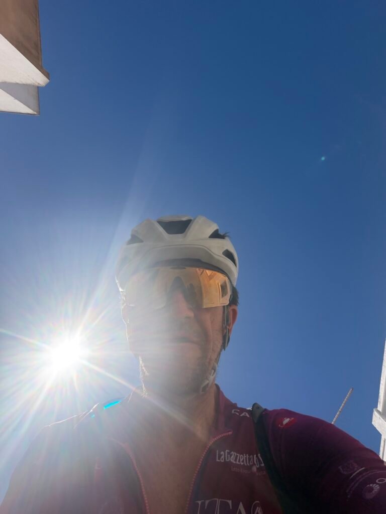 Richard in his Maglia Ciclamino jersey, white helmet and sunglasses taking a selfie against a deep blue sky with the sun flaring beside him, riding out of Alvor