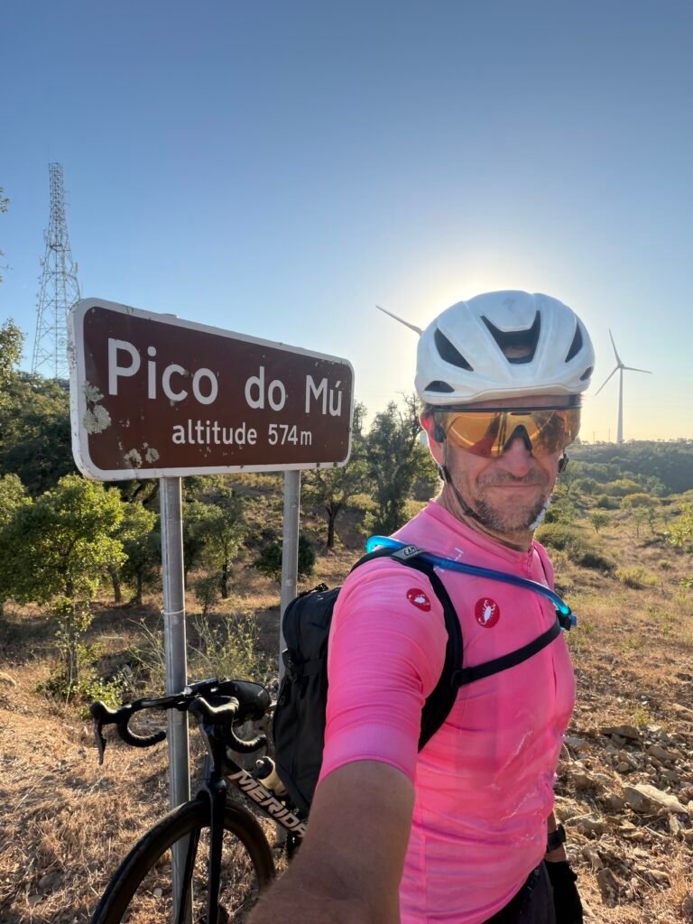 Richard in a pink Maglia Rosa jersey taking a selfie by the Pico do Mu sign, altitude 574 m, with a mast and a wind turbine behind in the evening sun