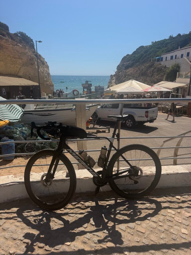 Black road bike leaning on a railing above Benagil beach, with café umbrellas and golden cliffs behind