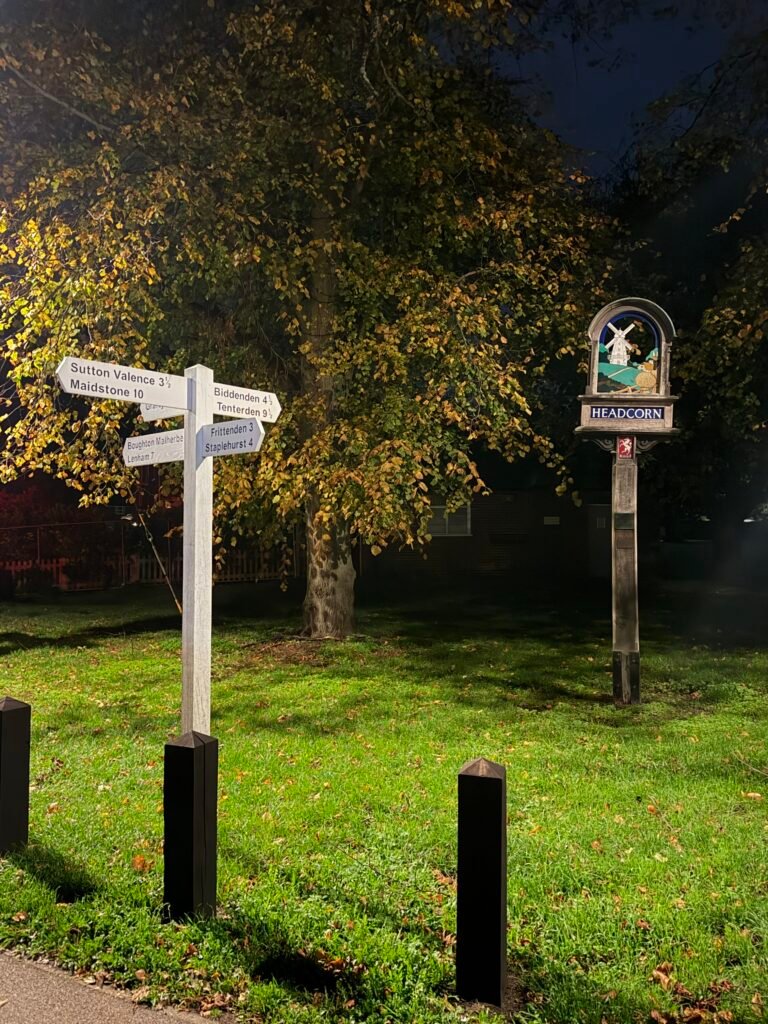 Headcorn village sign and a white fingerpost on the green, lit up at night under an autumn tree