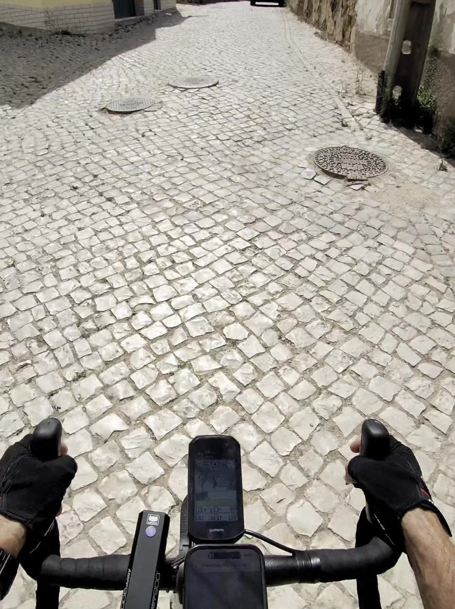 Rider's-eye view over the handlebars and Garmin down a narrow street of rough white cobbles with manhole covers, in Vila do Bispo