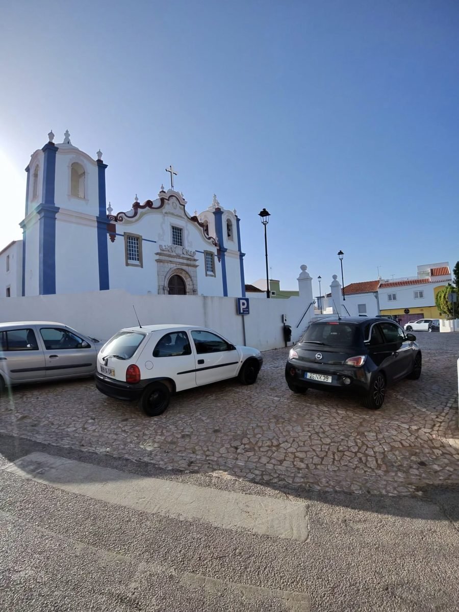 Whitewashed church with blue trim and a bell tower in Estombar, above a small car park, under a deep blue sky