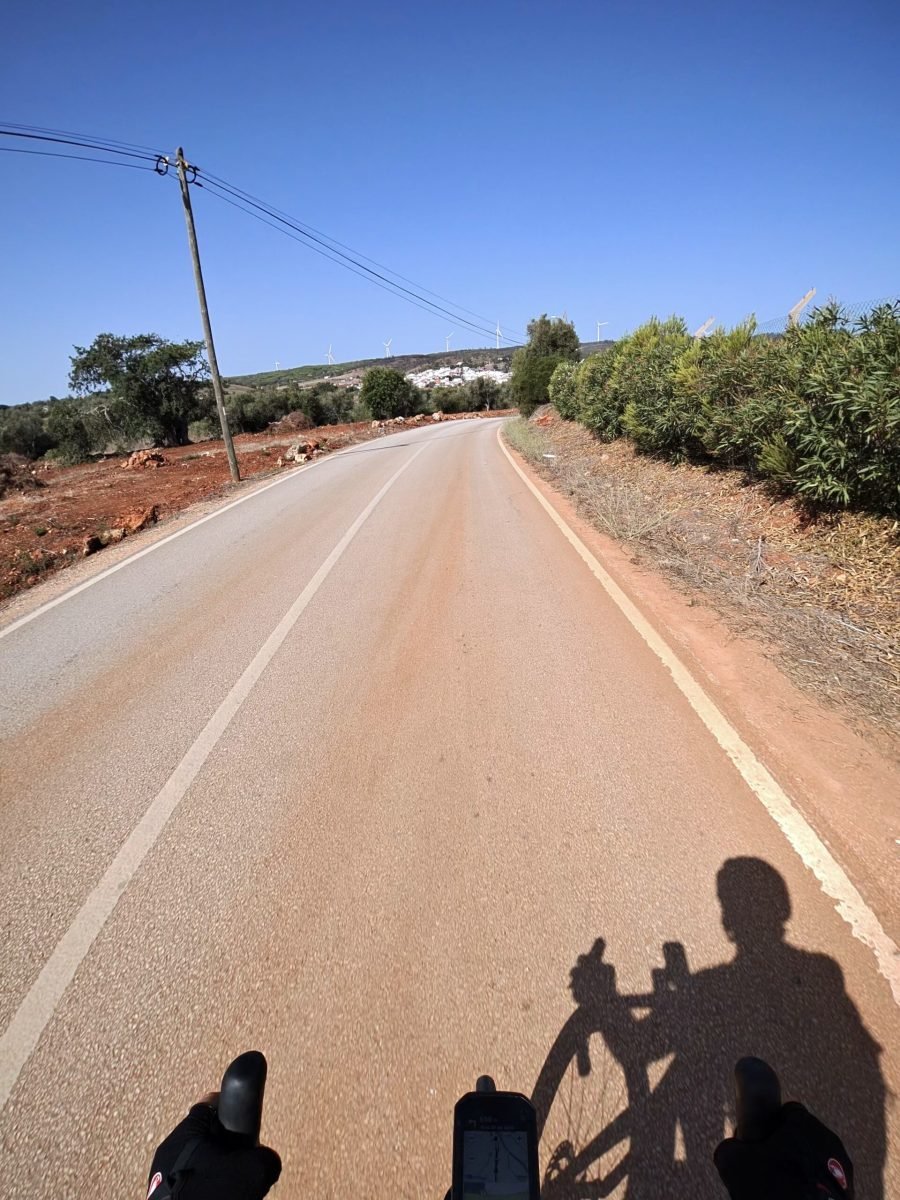 Handlebar view down an empty road with red earth verges and scrub, the rider's shadow on the tarmac