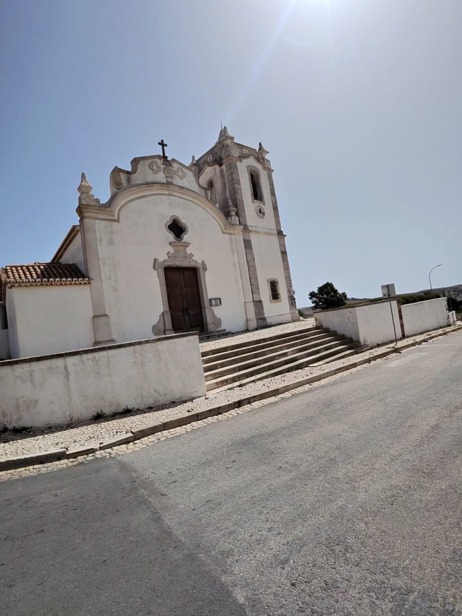 A whitewashed baroque church with a bell tower and steps up from a cobbled pavement under a bright sky