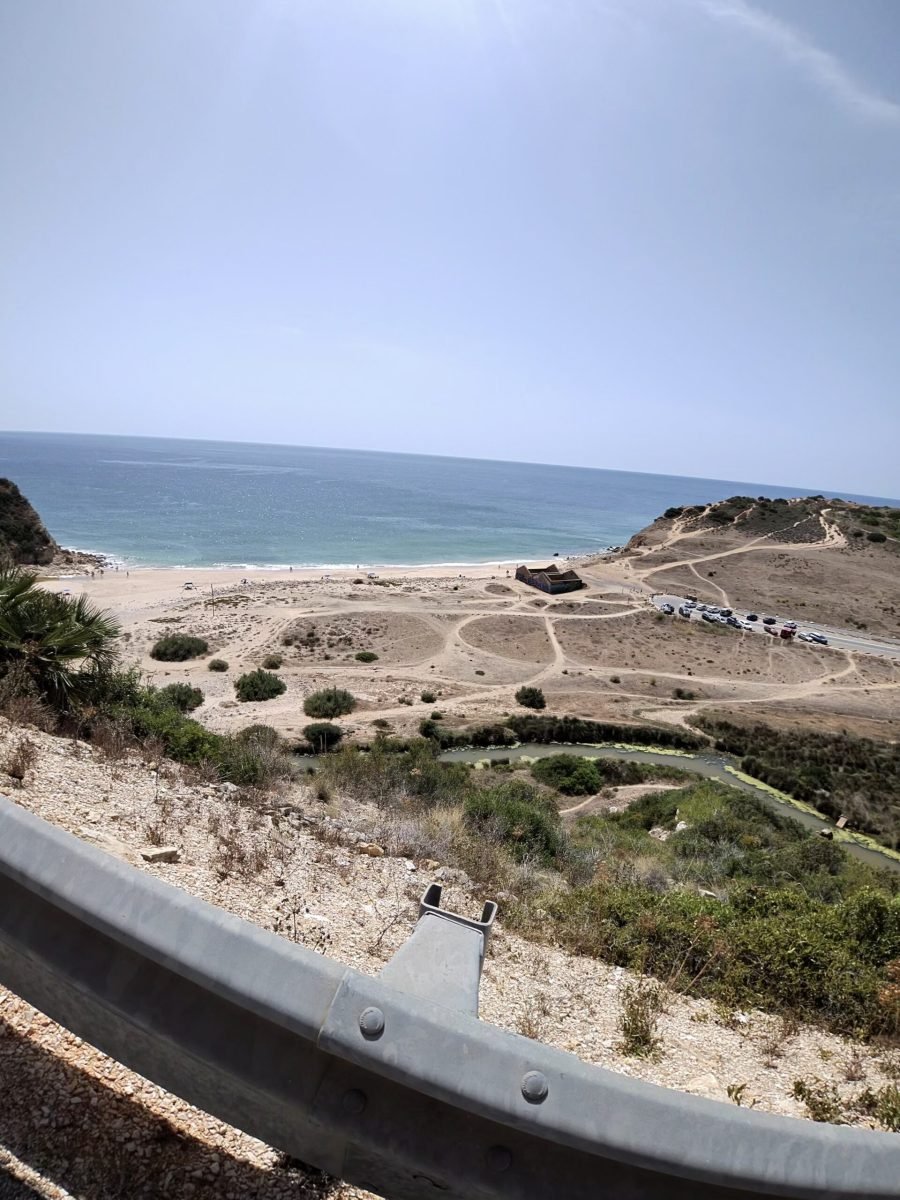 Looking down from the road over a crash barrier to a sandy beach and turquoise sea, with an old ruined building near the shore