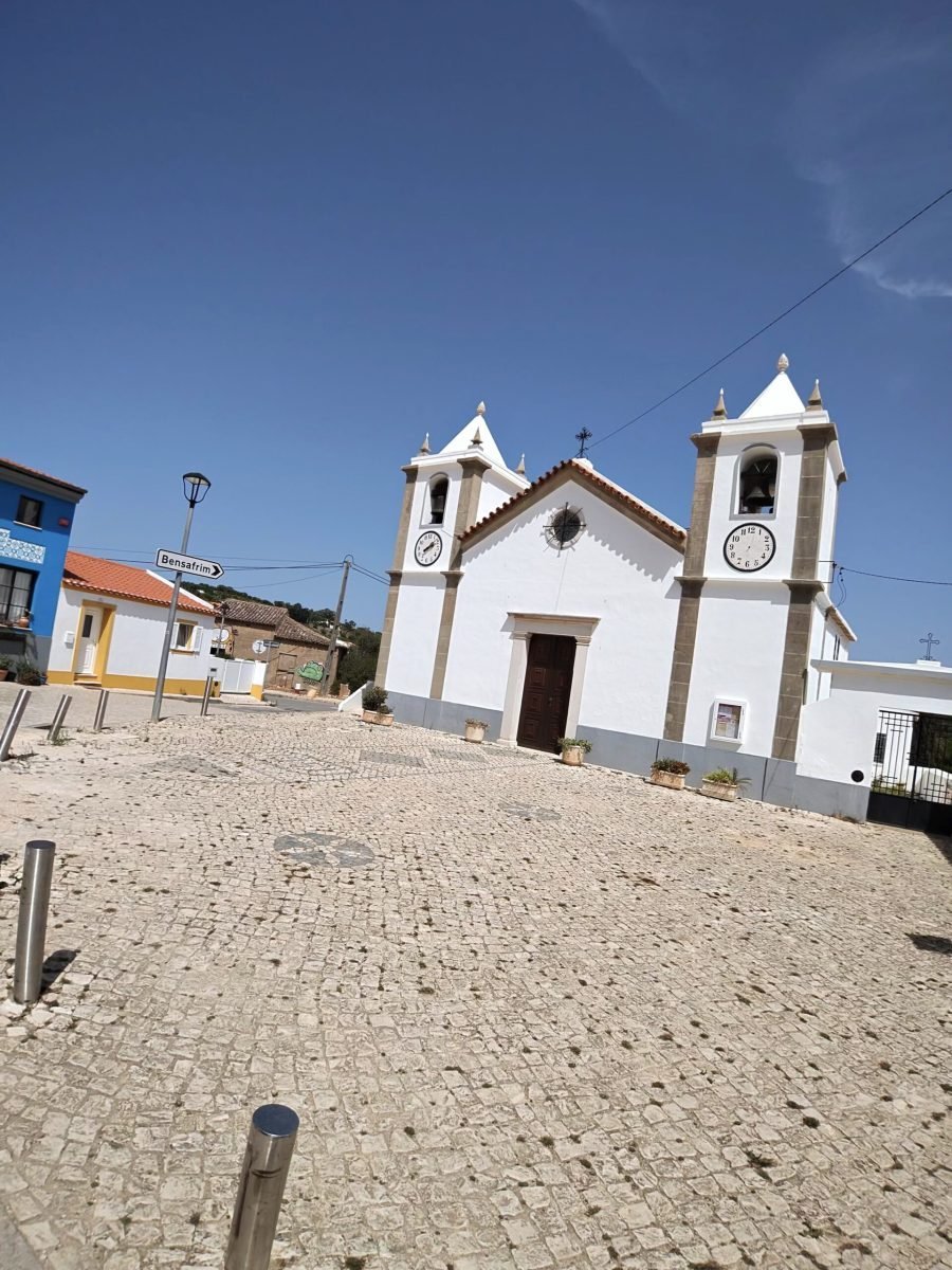 A white village church with twin clock towers behind a cobbled square, with a signpost to Bensafrim