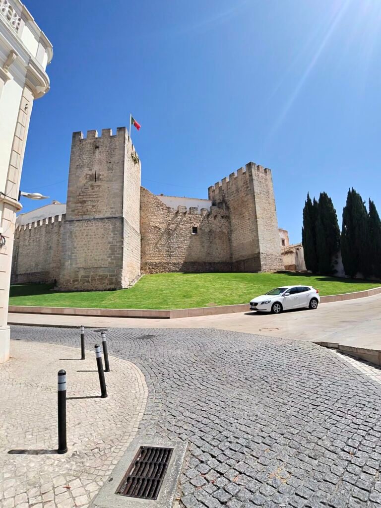 The stone towers and walls of Loulé castle above a cobbled street under a deep blue sky