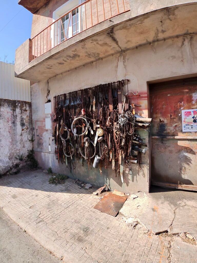 Rusty chainsaw chains and guide bars, with a few wheel rims and coils of cable, hung on the wall of a weathered workshop on the road to Alte