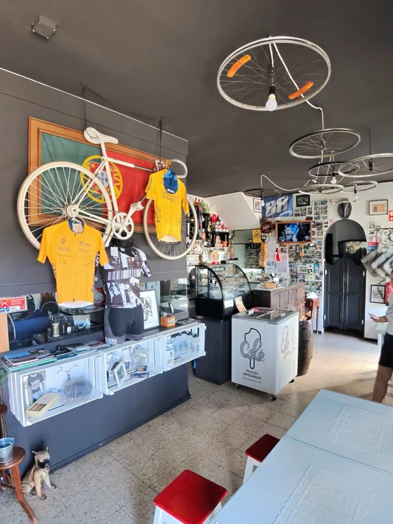 Inside the Germano biciArte Café in Alte: a white track bike mounted on a Portuguese flag, yellow jerseys, display counters and old bike wheels used as ceiling lights