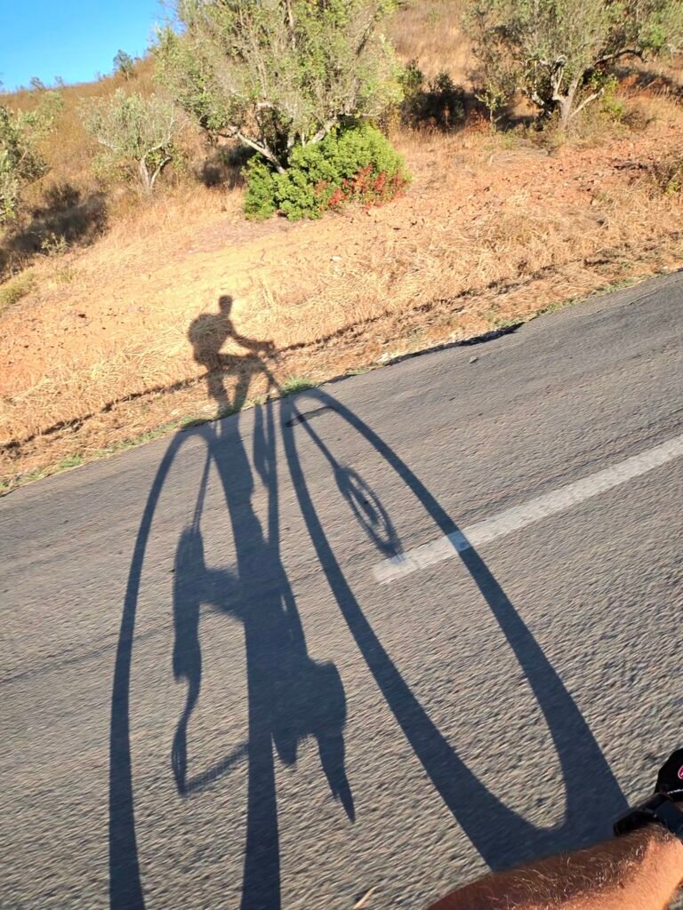 The rider's long shadow on the tarmac below a dry, scrubby hillside near the Arade Dam