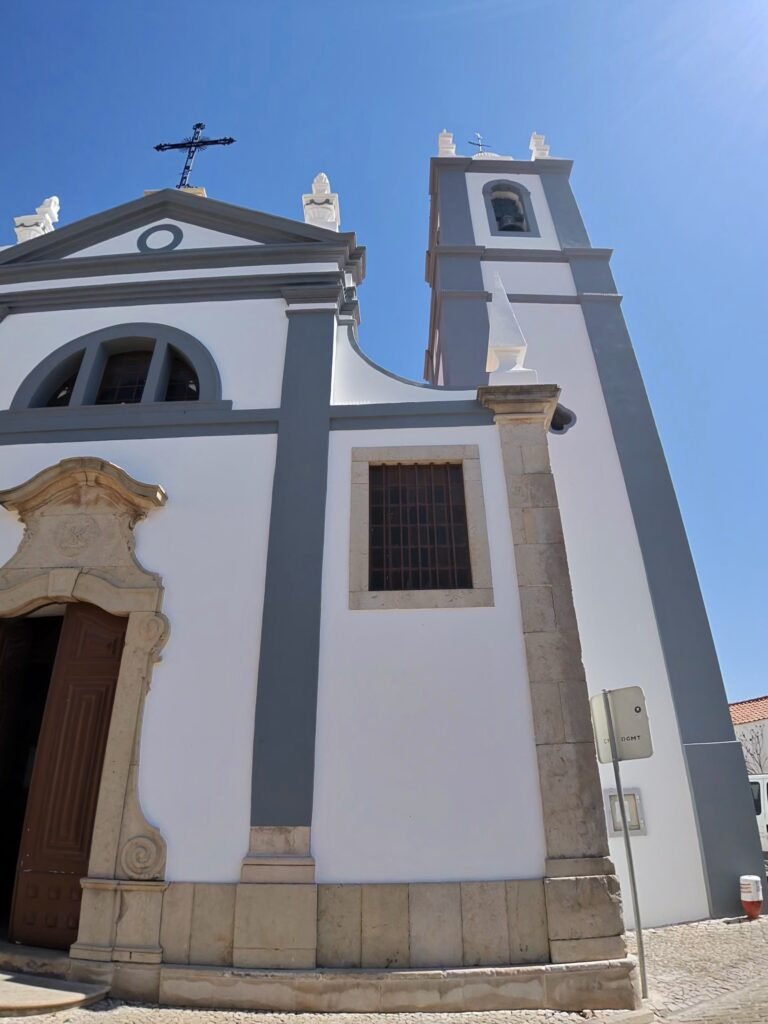 A white church with grey trim and a bell tower against a blue sky in Alcantarilha