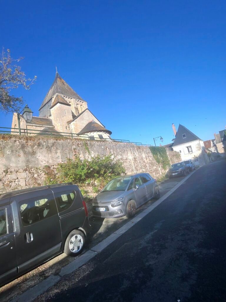 The church at Villandry above a stone wall, with cars parked along the climbing street under a blue sky