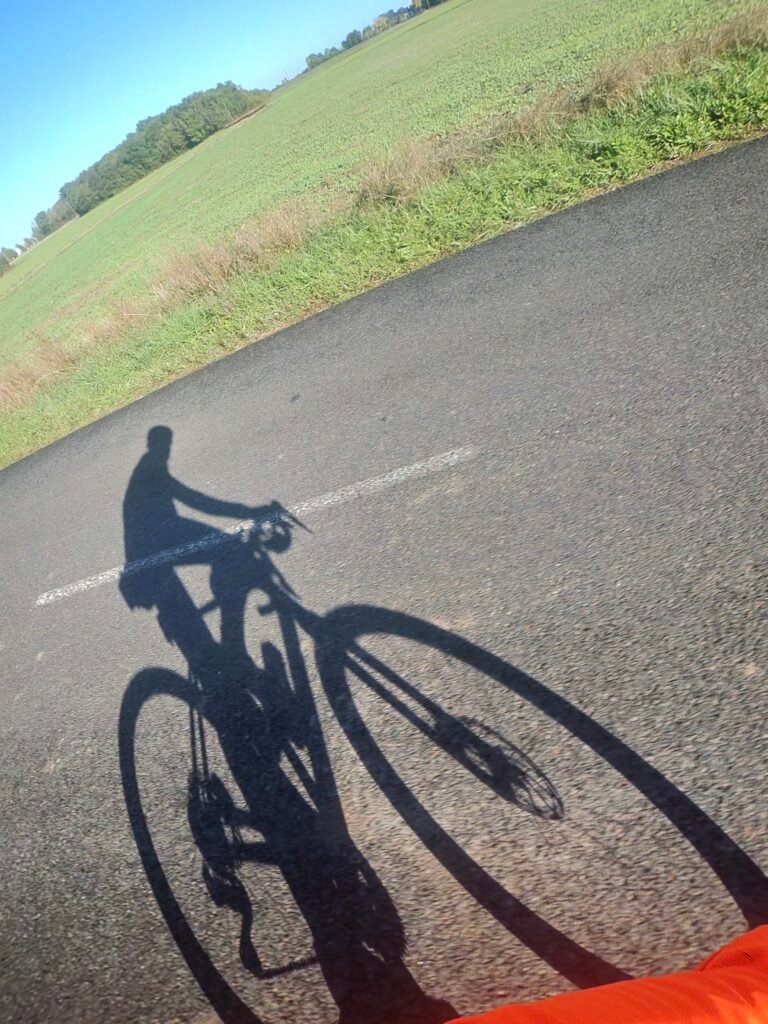 Richard’s shadow on the bike cast across the road beside a green field near Druye