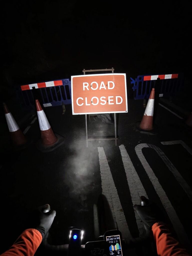 Rider's view over the handlebars at night: a Road Closed sign, barriers and cones in the headlight, breath fogging in the cold air