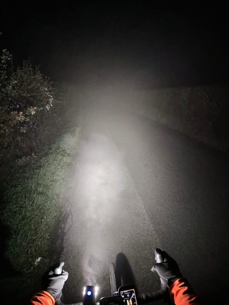 Rider's view over the handlebars at night: the headlight beam turned into a white cone by thick fog over a narrow lane with a grassy verge and hedge