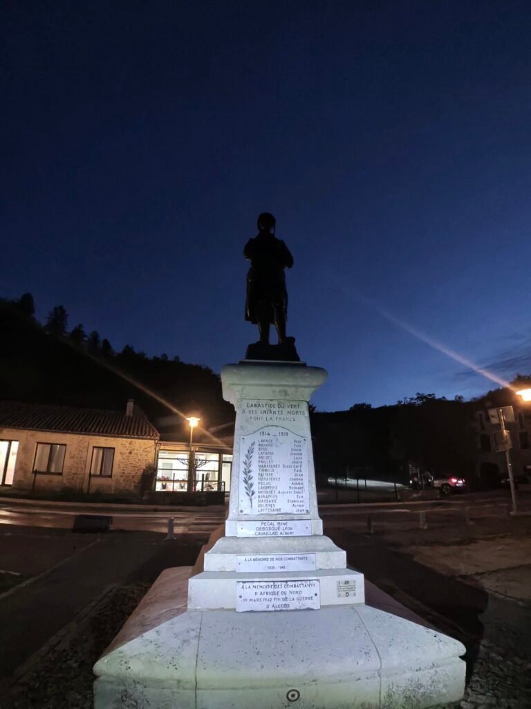A statue on a white stone plinth lit up against a deep-blue night sky in Labastide-du-Vert